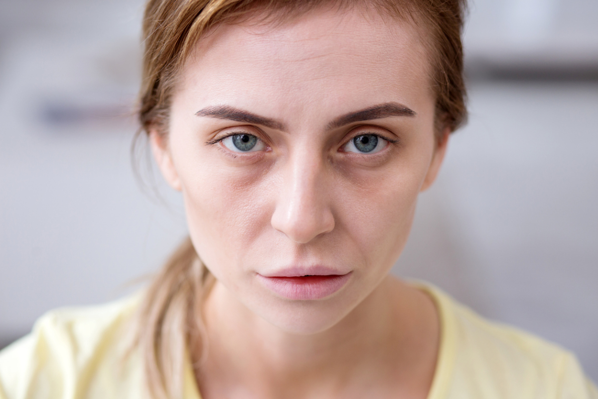 A woman with light blue eyes looks directly at the camera with a serious expression. She has light brown hair tied back and wears a pale yellow shirt.