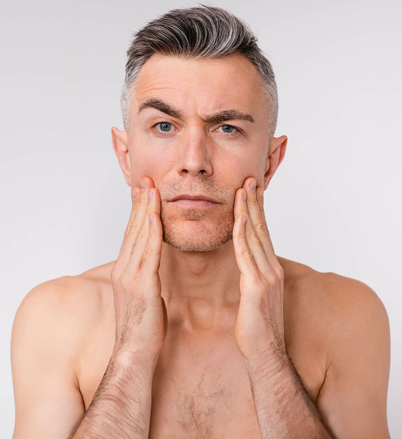 A shirtless man with short gray hair touches his cheeks with both hands. He has a concerned expression, examining his skin against a plain background.