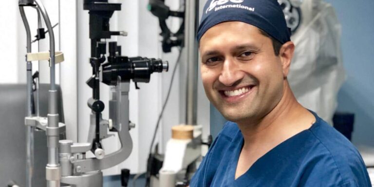 Smiling Dr. Parag Gandhi in blue scrubs and cap sits next to medical equipment in a clinical setting, conveying professionalism and warmth.