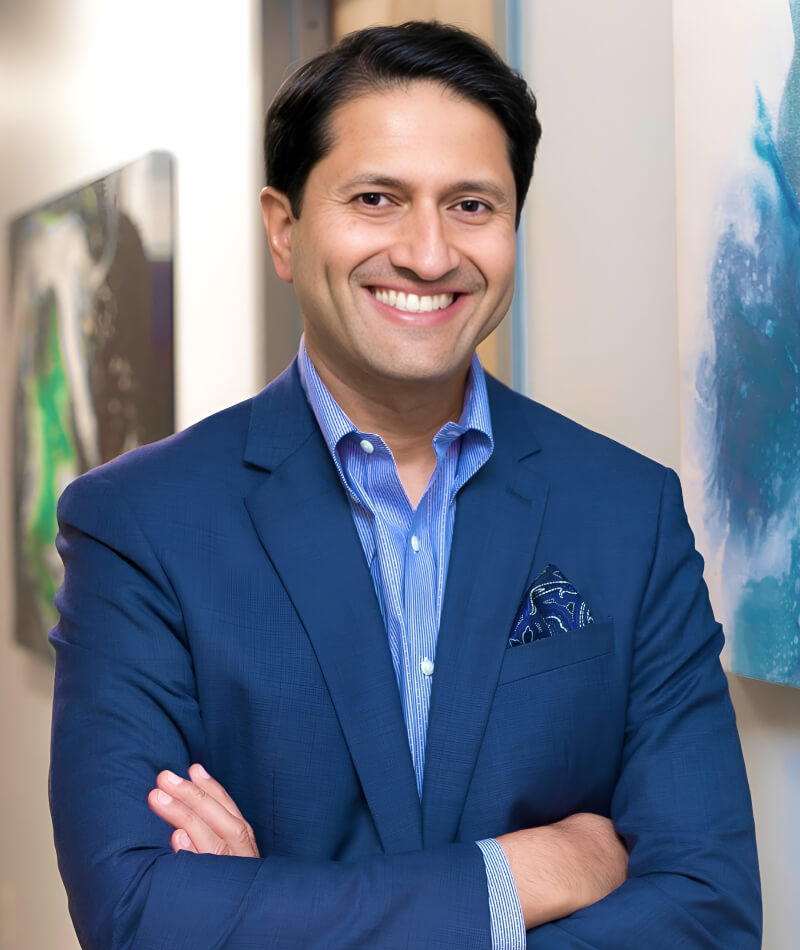 Dr. Parag Gandhi in a blue suit with crossed arms stands in a hallway with abstract art on the walls, conveying confidence and professionalism.