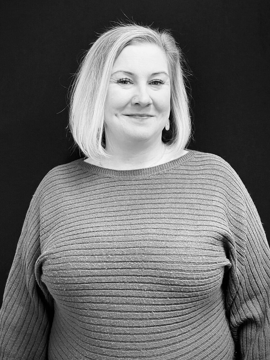 Black and white portrait of a woman with shoulder-length hair, smiling softly. She wears a textured sweater and stands against a dark background.
