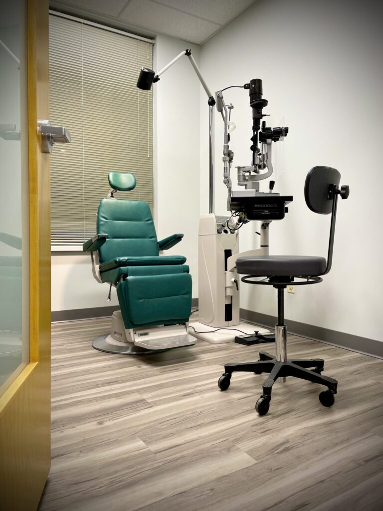 Optometrist exam room with a green reclining chair, slit lamp, and stool. The room has wood flooring and a window with closed blinds, creating a clinical atmosphere.