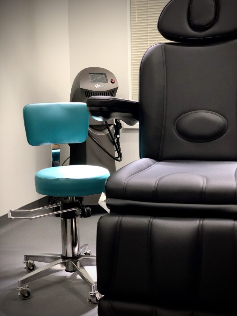 A modern medical room features a sleek black reclining chair and a turquoise stool with wheels. A laser machine stands in the background, conveying a calm, clinical atmosphere.