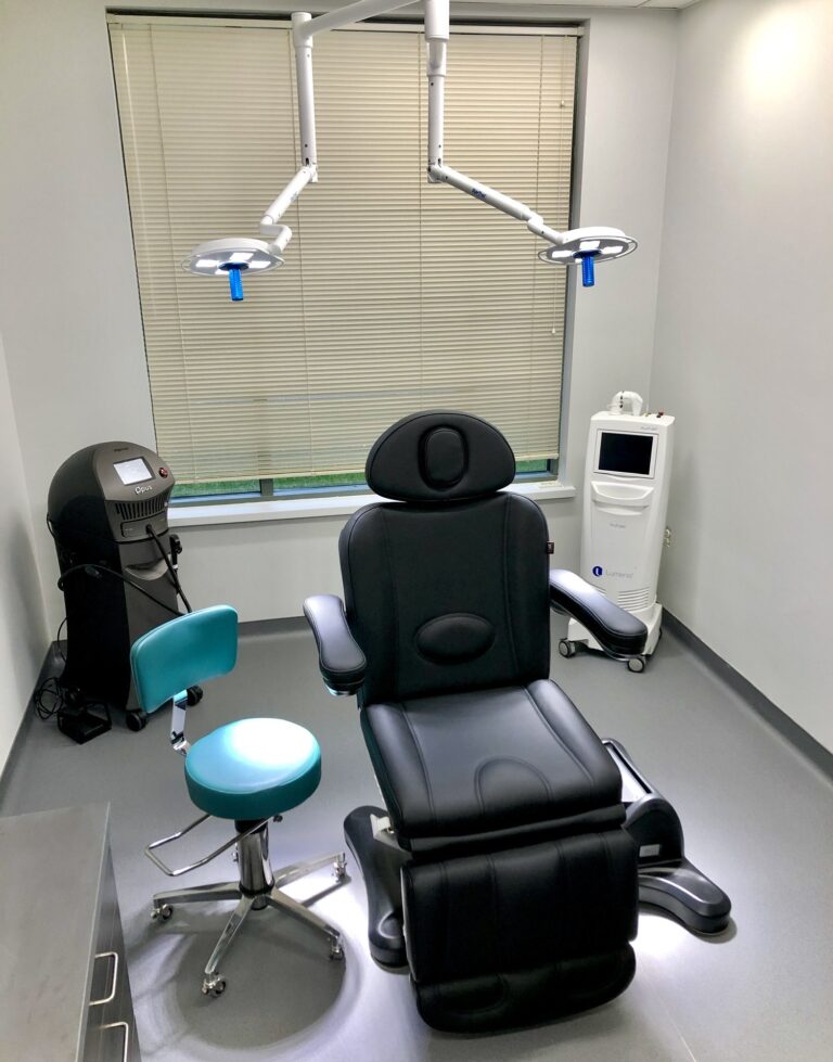 Modern medical examination room with a black reclining chair, turquoise stool, overhead lights, and equipment. The room is clean and orderly.
