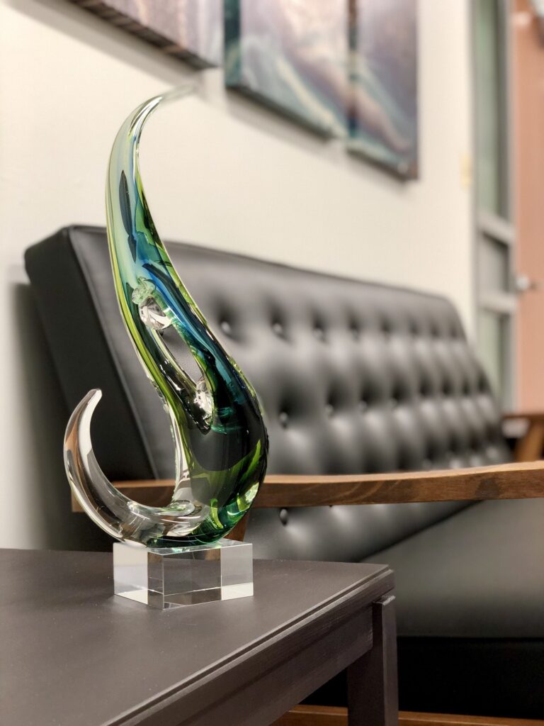 A sleek, glass sculpture with blue and green curves sits on a dark wooden table, against a tufted gray sofa. The setting feels modern and elegant.