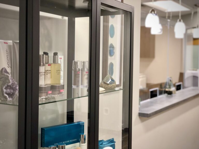Glass cabinet displaying skincare products and decorative stones, set in a modern interior with pendant lights and a sleek reception desk in the background.